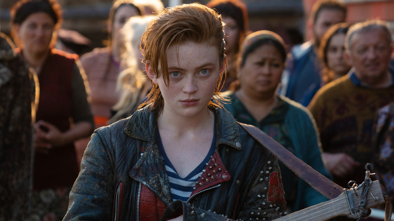 Sadie Sink as O'Dessa staring ahead with a guitar as a crowd stands behind her in O'Dessa