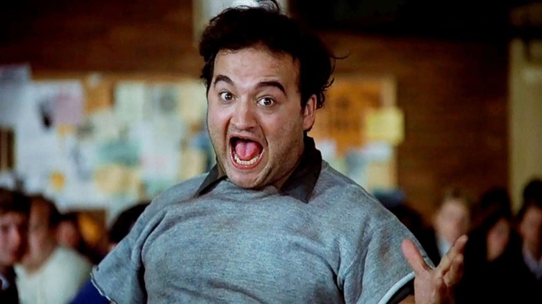 John Belushi as John "Bluto" Blutarsky in Animal House, with his hands raised and mouth open wide while looking at the camera.