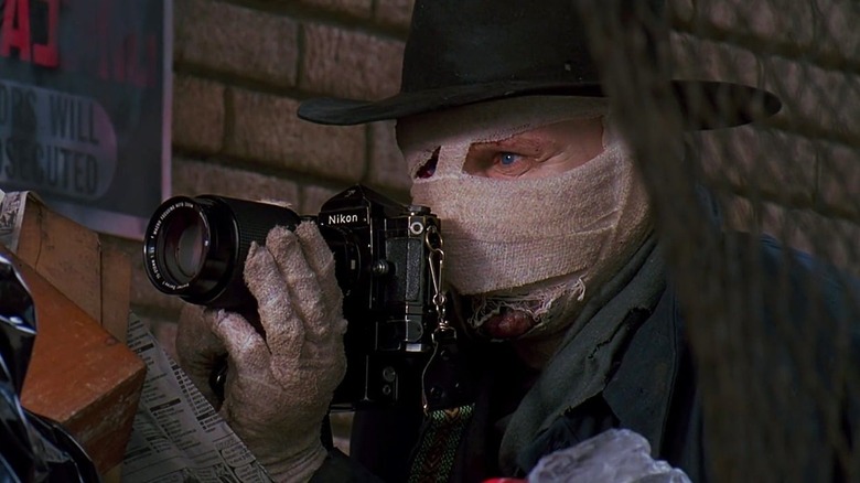 Liam Neeson as Dr. Peyton Westlake in Darkman, with a bandaged face and wide-brim hat on as he looks over his camera while taking photos of something.