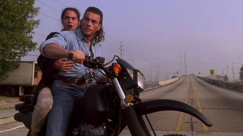 Jean-Claude Van Damme as Chance Boudreaux and Yancy Butler as Natasha Binder in Hard Target, with Natasha holding on Chance on the back of a motorbike in front of an empty highway bridge.