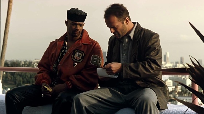 Bruce Willis as Joe Hallenback and Damon Wayans as Jimmy Dix in The Last Boy Scout, sitting next to each other against the skyline and investigating something in Joe's hands.