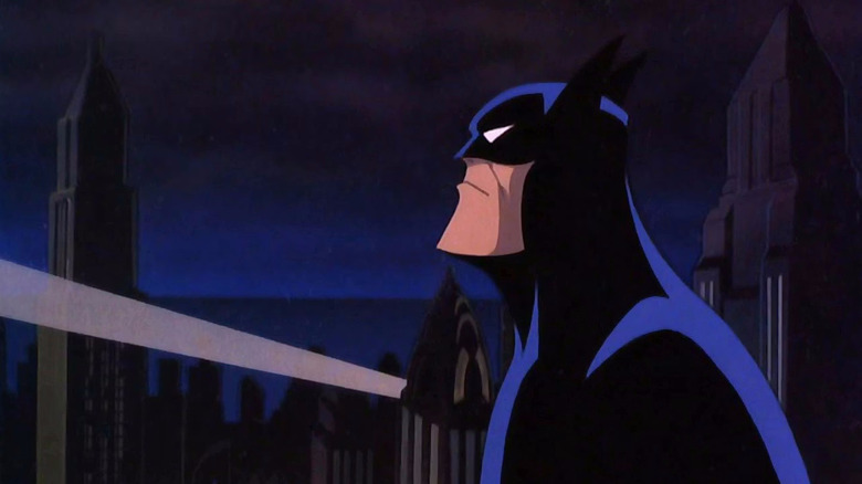 Batman (voiced by Kevin Conroy) in Batman: Mask of the Phantasm, seen in profile as he stands on a rooftop with the bat signal shining off within the Gotham cityscape behind him.