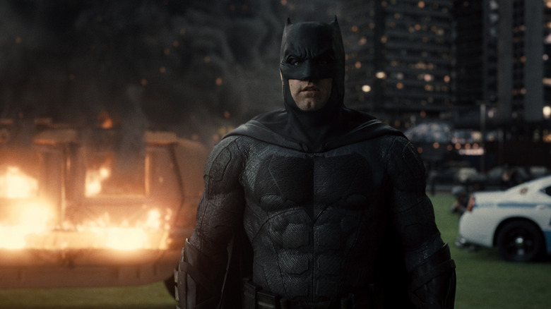 Ben Affleck as Bruce Wayne/Batman in Zack Snyder's Justice League, standing in his Batsuit, with a burning helicopter in the background.