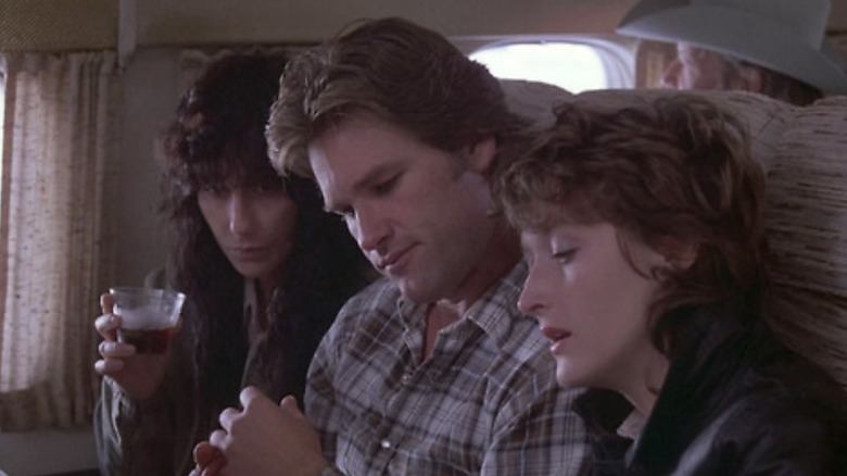 Cher as Dolly Pelliker holding a drink, Kurt Russell as Drew Stephens, and Meryl Streep as Karen Silkwood sitting together on a place while reading in Silkwood