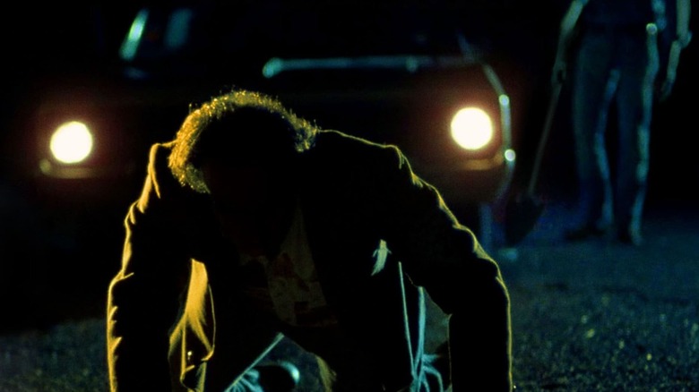 Julian Marty (Dan Hedaya) kneels before a car with its headlights on as Loren Visser (M. Emmet Walsh) walks up holding a shovel in Blood Simple