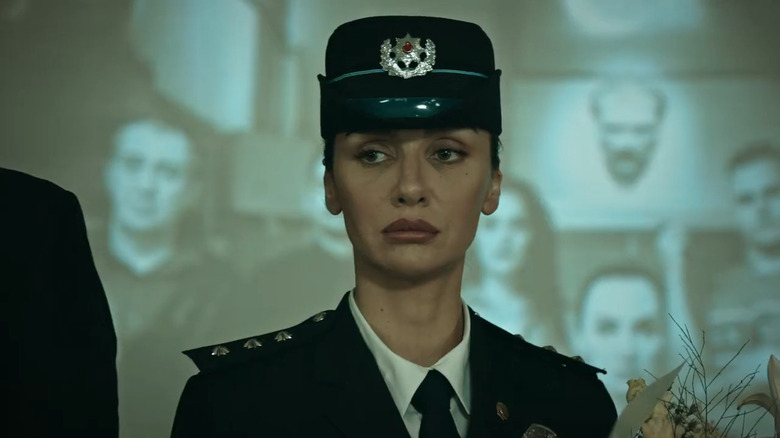 Birce Akalay as Önem Özülkü, in dress police uniform and hat holding flowers with image projected behind her in Graveyard