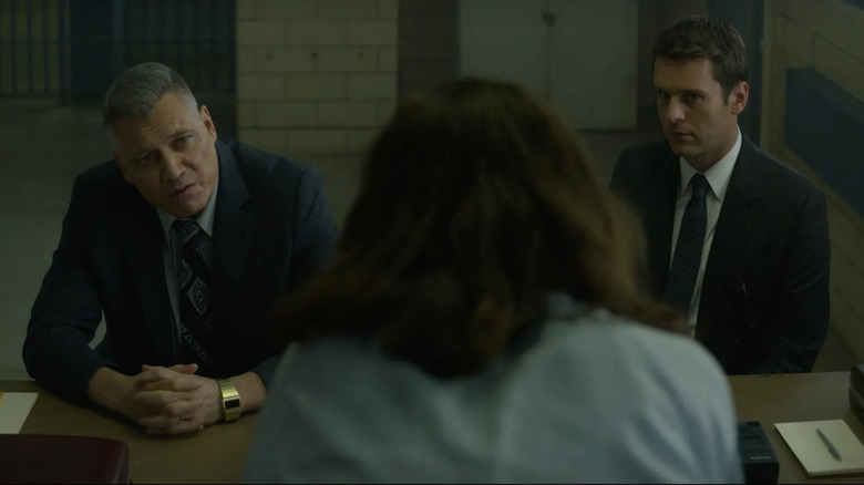 Holt McCallany as Bill Tench sitting next to Jonathan Groff as Holden Ford at table sitting across from long-haired man (face not pictured) in Mindhunter