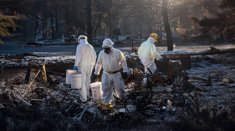 Three people in white jumpsuits, hard hats, and masks cleaning up debris in burnt forest in Fire in Paradise