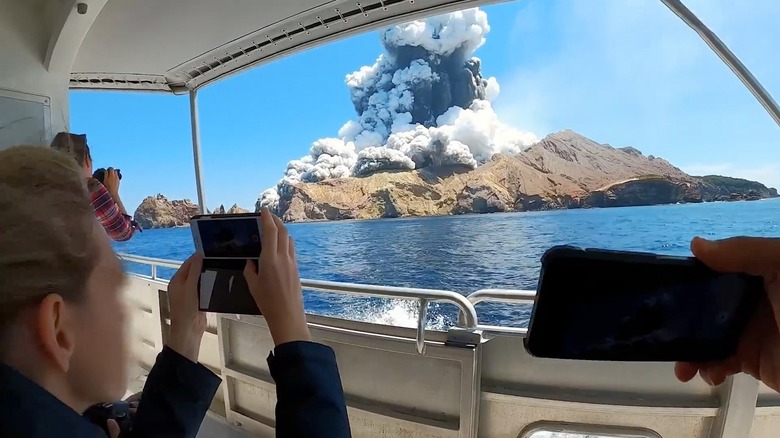 Several people (faces not visible) recording volcano exploding on phones and cameras from boat in The Volcano: Rescue From Whakaari