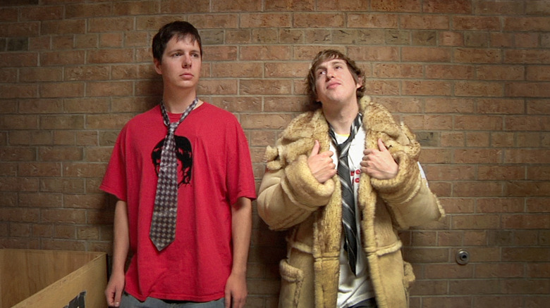 Matt Johnson as Matt and Owen Williams as Owen in The Dirties, with Matt in a fur coat and Own in a t-shirt and a loose tie as they stand against the brick wall of the interior of their high school.