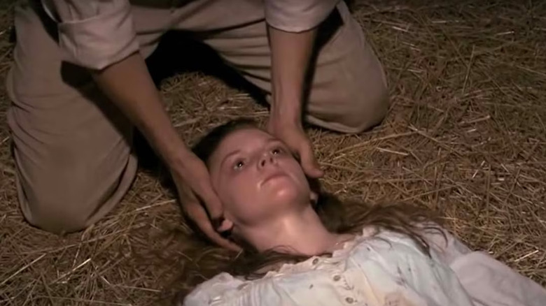Ashley Bell as Nell Margaret Sweetzer in The Last Exorcism, laying on the hay-strewn ground and looking straight above her as a man kneels over her head and holds her head with both hands.