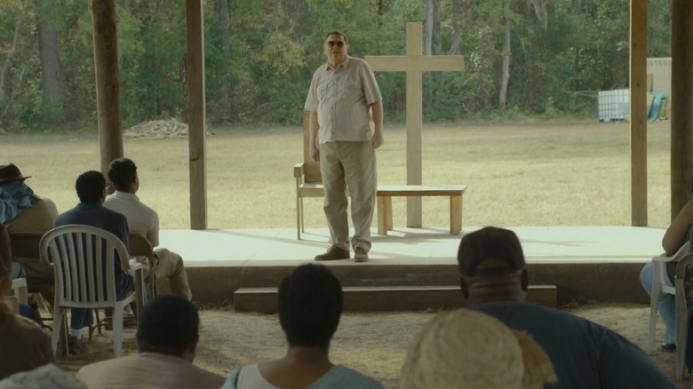 Gene Jones as Father in The Sacrament, standing at his makeshift pulpit with a wooden cross behind him as he makes his sermon to the crowd of cult-followers.