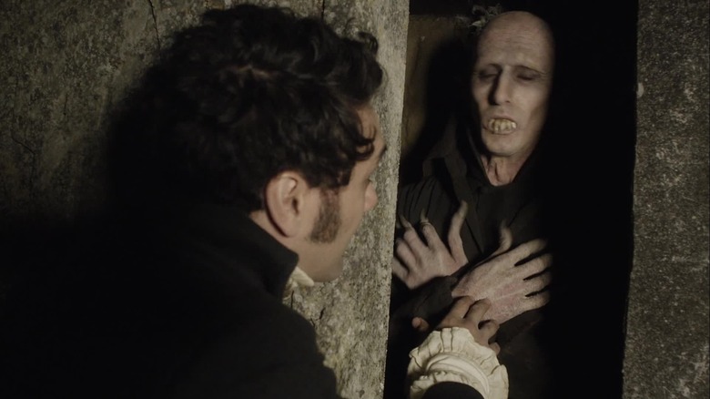 Taika Waititi as Viago tapping on the hand of Ben Fransham as Peytr, who is a Nosferatu-style vampire sleeping in a stone coffin, in What We Do in the Shadows