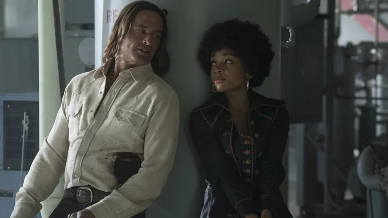 Josh Holloway as Jim Ellis and Rachel Hilson as Nina Hayes leaning against a pillar and holding guns in Duster