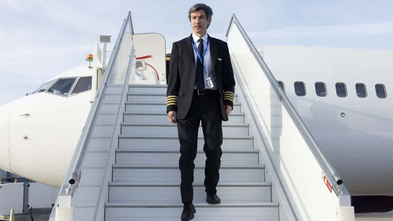 Nathan Fielder in a pilot uniform descending the steps of an airplane