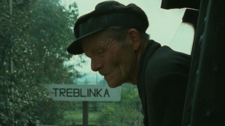 A man peers out of a train passing a sign reading Treblinka in Shoah