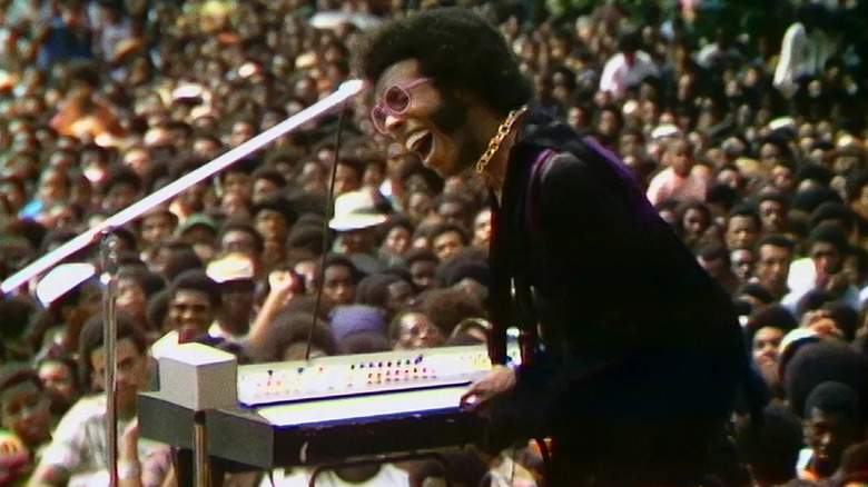 Sly Stone jubilantly plays a keyboard on stage before a large crowd in Summer of Soul