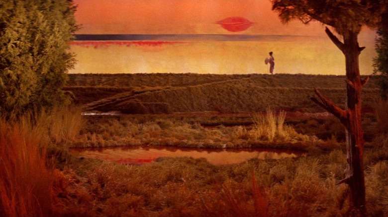 One of the painterly vistas found within Kwaidan, a red and orange-tinged landscape among the grassy hills with someone standing at a distance.