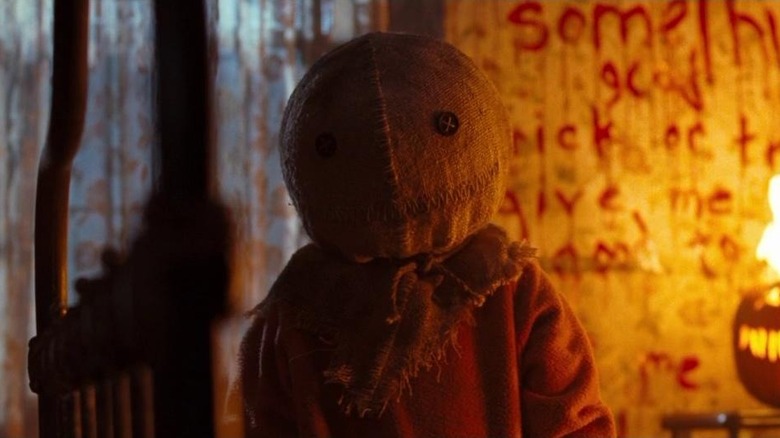 Quinn Lord as Sam in Trick 'r Treat, looking directly on in his creepy burlap sack mask, with scary scribblings on the wall behind him, along with a burning Jack-o'-lantern.