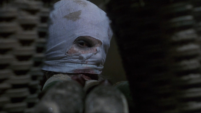 Jonathan Fuller as Giorgio Orsino, The Freak, in Castle Freak, seen through some kind of structure surrounding his head, which is covered in makeshift bandages as he looks on.