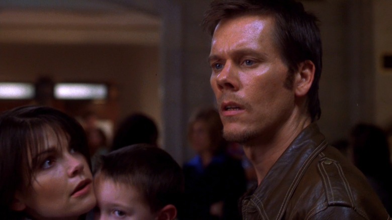 Kevin Bacon as Tom Witzky in Stir of Echoes, looking slightly concerned over at something while Kathryn Erbe as Maggie holds Zachary David Cope as Jake and looks up at him from the side.