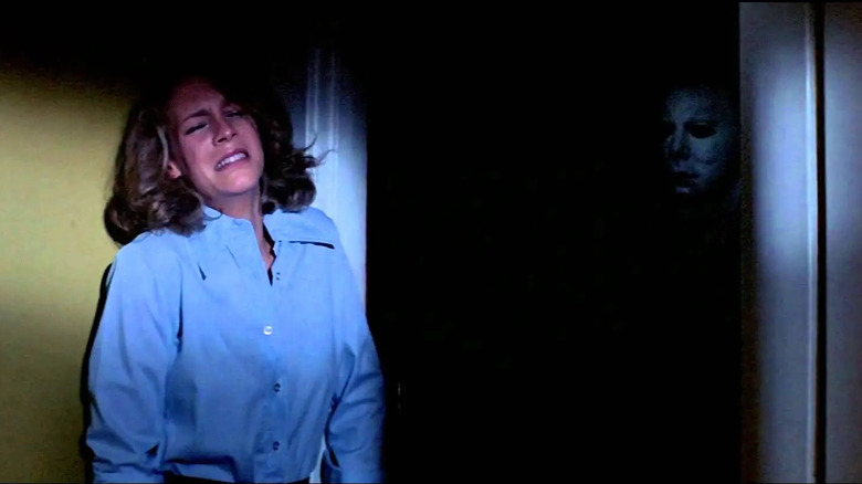 Jamie Lee Curtis as Laurie Strode in Halloween, leaning against the wall and crying while Nick Castle as Michael Myers peers at her from the pitch-black dark of the next room.