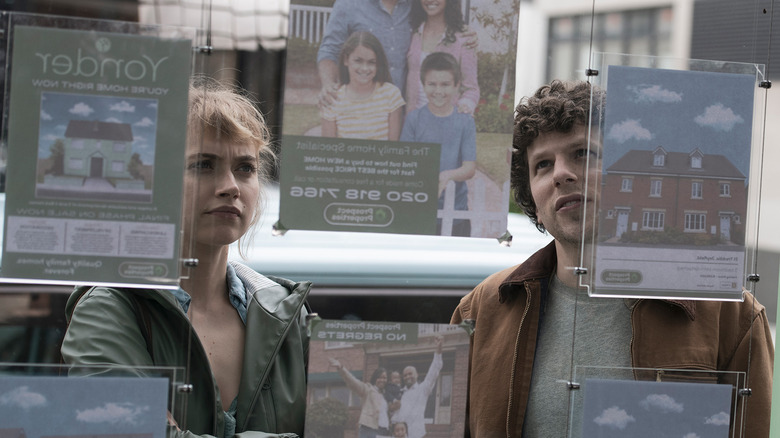 Imgoen Poots as Gemma and Jesse Eisenberg as Tom looking at the window of a real estate firm with signs on it for available properties in Vivarium