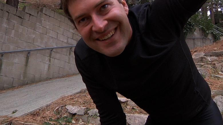 Mark Duplass as Josef in Creep, leaned over and smiling while he looks at whoever is above the frame of the camera.