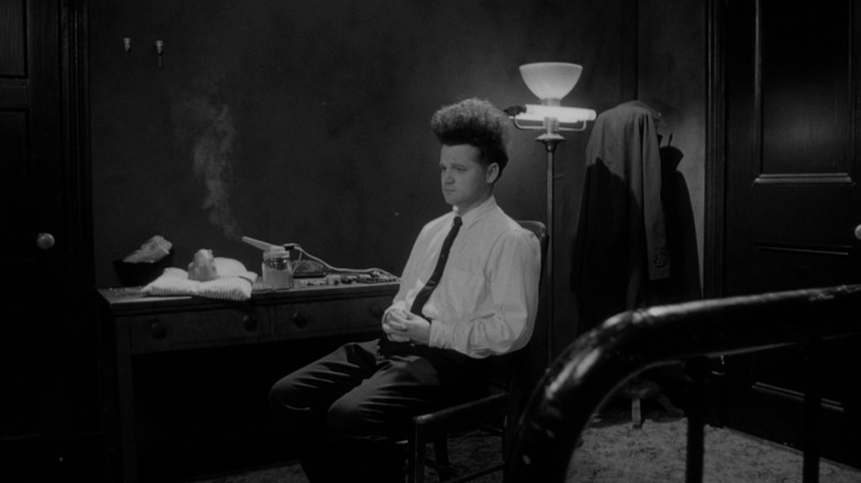 Jack Nance as Henry Spencer in Eraserhead, sitting in his sparse apartment with the bundled up mutant infant on the desk behind him as he sits in the middle of the room alone.