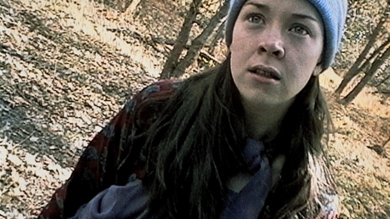 Heather Donahue as a fictionalized version of herself in The Blair Witch Project, standing in the woods close to the slanted camera in her jacket and beanie.