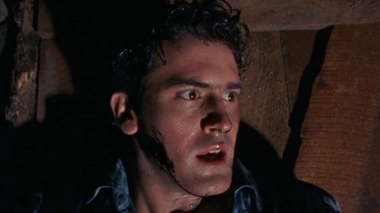 Bruce Campbell as Ash Williams in The Evil Dead, sweating and looking anxious as he stands against a wooden door.