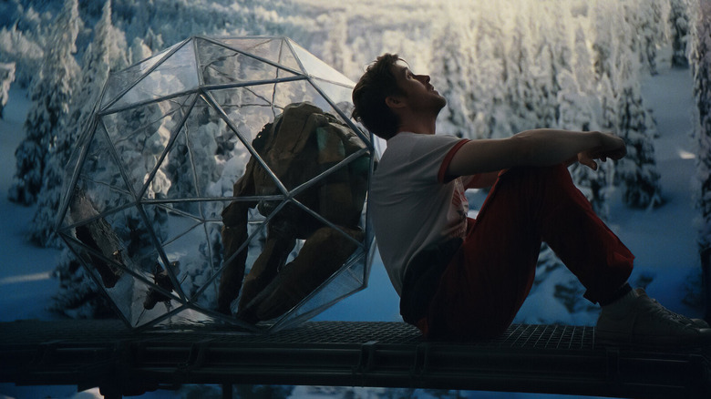 Ryan Gosling as Ryland Grace, leaning up against the transparent shell housing Rocky, puppeted and voiced by James Ortiz, as the pair sit in front of a projection of a snowy landscape.