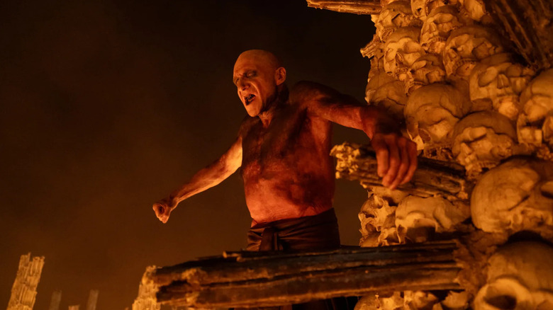 Ralph Finnes as Dr. Ian Kelson in 28 Years Later: The Bone Temple, covered in iodine and raising his fist as he perches among his Bone Temple