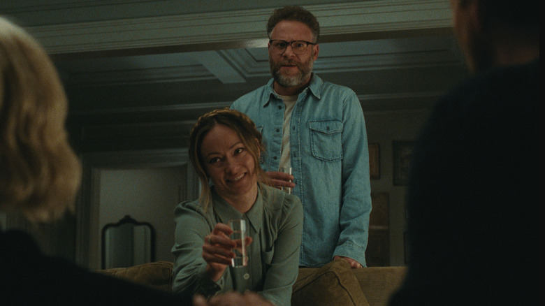 Seth Rogen as Joe in The Invite, standing behind Olivia Wilde as Angela sitting on a couch and holding a glass.