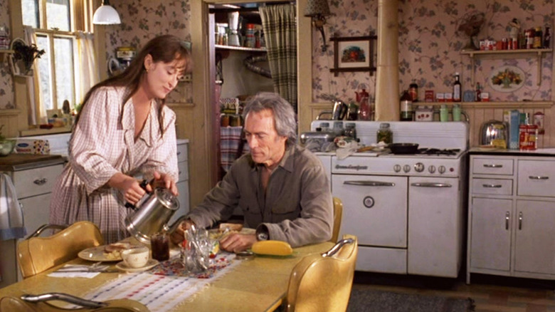 Clint Eastwood as Robert Kincaid and Meryl Streep as Francesca Johnson in The Bridges of Madison County, together in a homey kitchen with Robert sitting at the table and Francesca pouring him coffee.