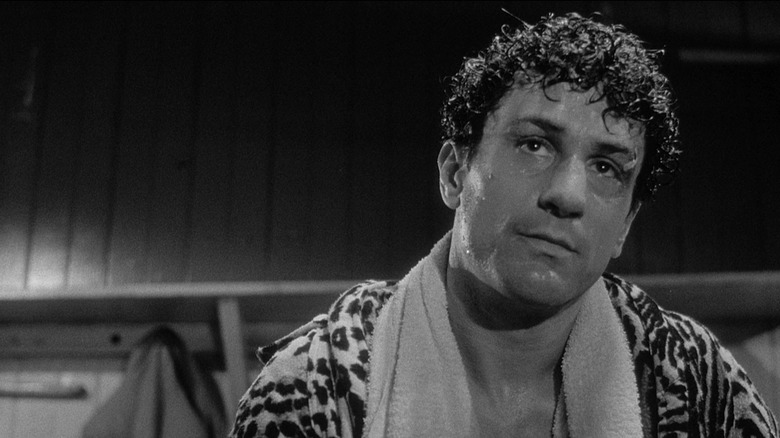Robert De Niro as Jake LaMotta in Raging Bull, looking sweaty and bruised in a robe with a towel around his neck as he looks on at something.