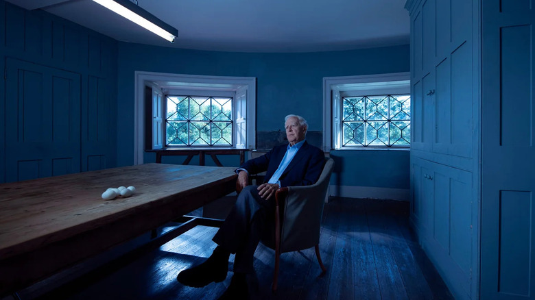 John le Carré, sitting cross-legged in a chair at a table.