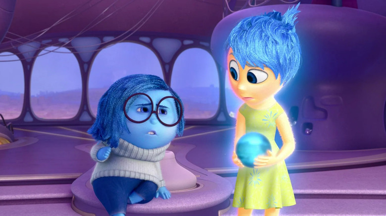 Animated Sadness, with blue skin, hair, and pants, sitting next to Happiness, standing in green dress and blue hair holding blue orb in Inside Out