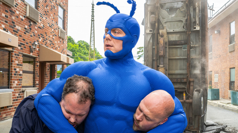 The Tick (Peter Serafinowicz) with two bad guys in headlocks on The Tick