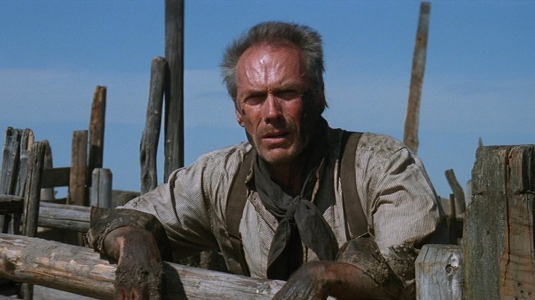 Clint Eastwood as William Munny in Unforgiven, looking dirty and tired and he stands in the sun and quints, leaning on a wooden pen.
