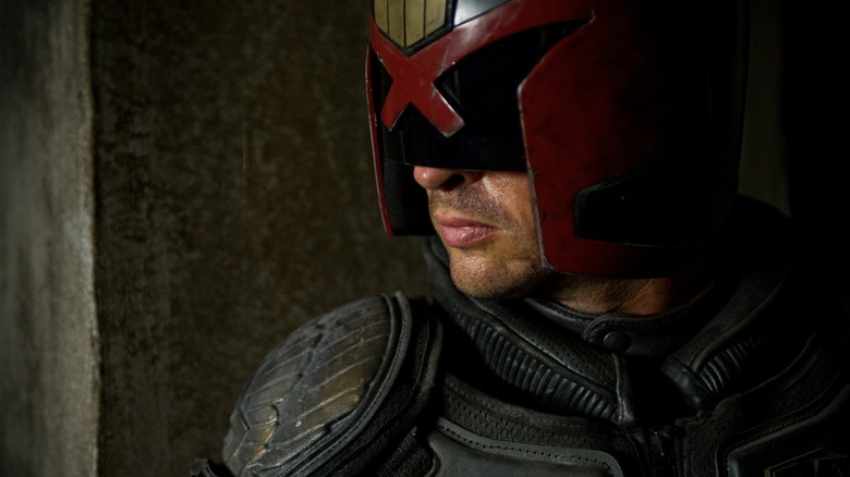 Karl Urban as Judge Dredd wearing his helmet and standing in front of a concrete wall in Dredd