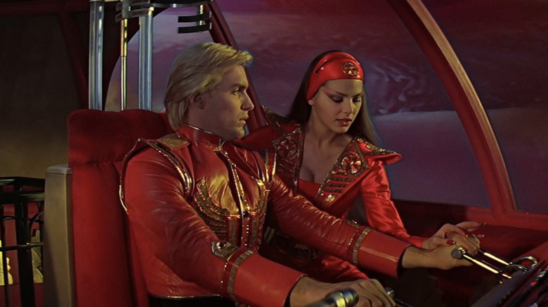 Sam J. Jones as Flash Gordon wearing a red jacket piloting a spaceship alongside Ornella Muti as Princess Aura wearing a red headband in Flash Gordon