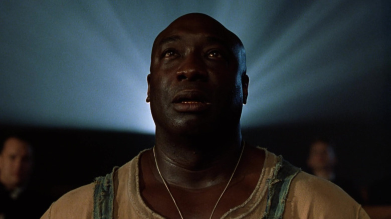Michael Clarke Duncan as John Coffey in The Green Mile, sitting with the glow of a projector surrounding his head, as he looks on at the screen out of frame.