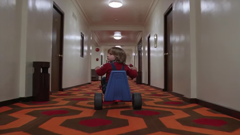 Danny Lloyd as Danny Torrance in The Shining, riding his tricycle through the iconic halls of The Overlook Hotel, turning his head back to look behind him.