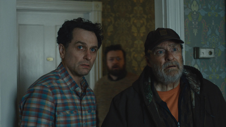 Matthew Rhys as Tom Loftis and Stephen Root as Wyck Crawford in Widow's Bay, standing next to each other with apprehension in a house, with Neil Casey as Kurt standing behind them