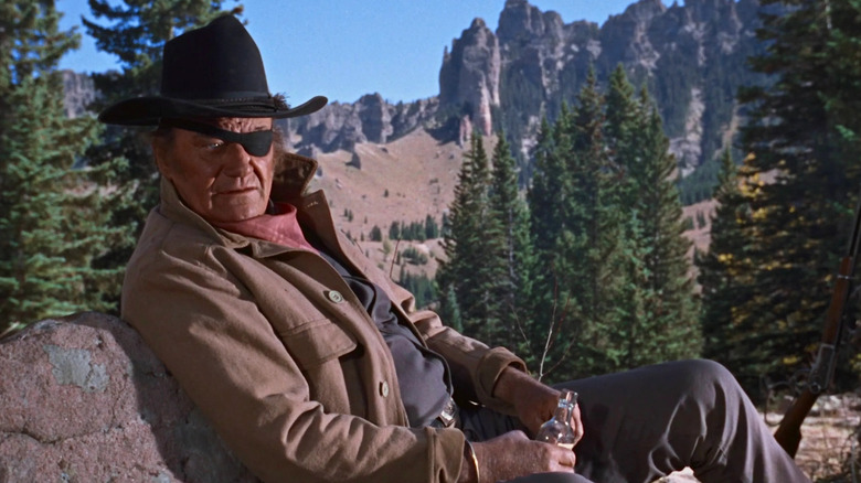 Rooster Cogburn (John Wayne) sits while holding a whiskey bottle in True Grit (1969)
