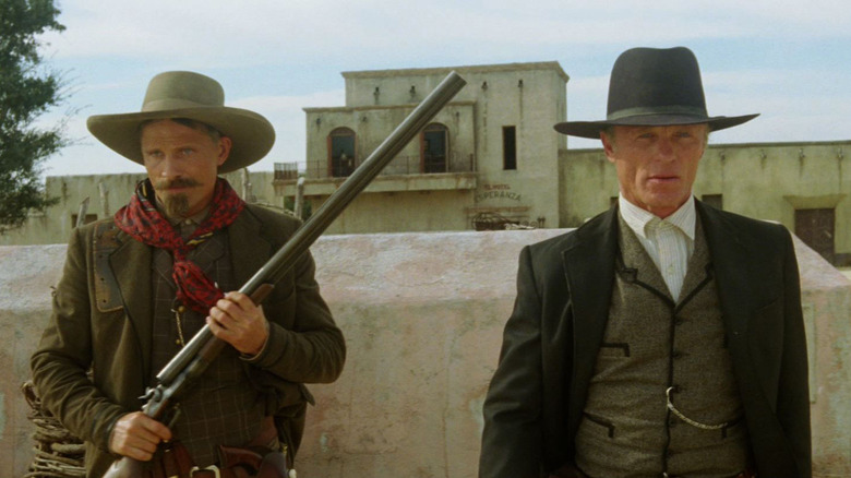 Viggo Mortensen as Everett Hitch and Ed Harris as Virgil Cole in Appaloosa, standing in front of a mission next to each other while Everett wields a rifle.