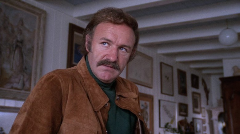 Gene Hackman as Harry Moseby in Night Moves, wearing a tan jacket in a room with a lot of art on the walls, looking out at something.