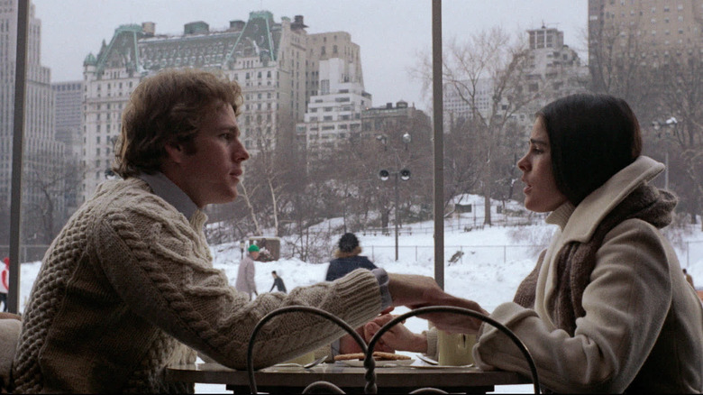 Ali MacGraw as Jennifer "Jenny" Cavilleri and Ryan O'Neal as Oliver Barrett IV in Love Story, sitting across from each other at a table in front of a snowy city backdrop.