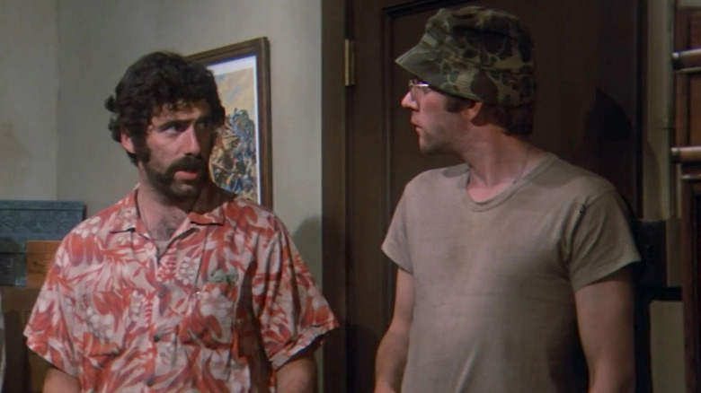 Donald Sutherland as Captain Benjamin Franklin "Hawkeye" Pierce Jr. and Elliott Gould as Captain John Francis "Trapper John" McIntyre in M*A*S*H, looking at each other, Sutherland in a camo army hat and Gould in a Hawaiin shirt.